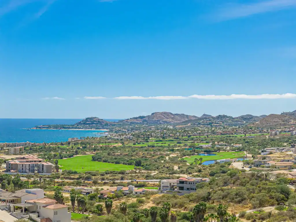 Aerial view of Club Campestre with Jack Nicklaus golf course fairways, residential neighborhoods, and the Sea of Cortez coastline extending toward Palmilla Point with Sierra de la Laguna foothills in the background